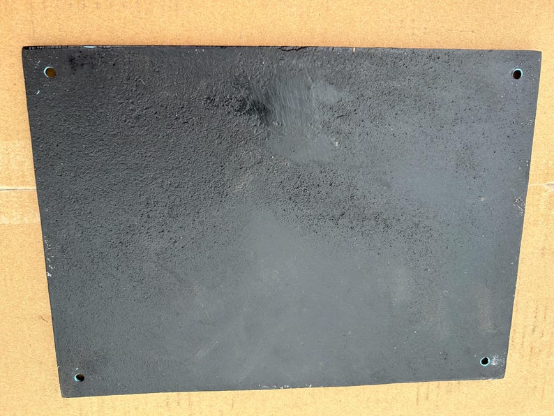 May include: A rectangular black metal plaque with a textured, matte finish. The plate features four small, circular mounting holes positioned near each of the four corners, allowing for secure attachment to a flat surface or wall structure.