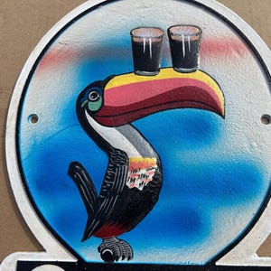 Puede incluir: Un letrero met&aacute;lico circular que presenta un tuc&aacute;n pintado con un pico grande y multicolor que equilibra dos vasos oscuros de cerveza stout sobre su pico. El fondo es un degradado de azul y blanco, con dos orificios para montaje.