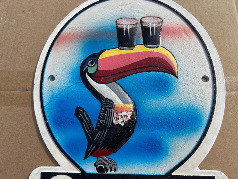 Puede incluir: Un letrero met&aacute;lico circular que presenta un tuc&aacute;n pintado con un pico grande y multicolor que equilibra dos vasos oscuros de cerveza stout sobre su pico. El fondo es un degradado de azul y blanco, con dos orificios para montaje.