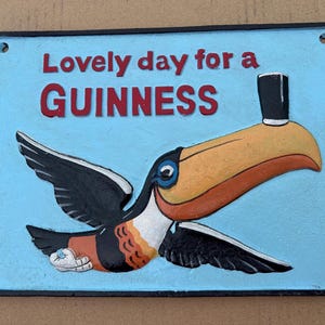 May include: A rectangular metal sign with a light blue background featuring a toucan with a black top hat on its beak. The text reads "Lovely day for a GUINNESS" in red letters. The toucan has black wings, a yellow beak, and a white chest.