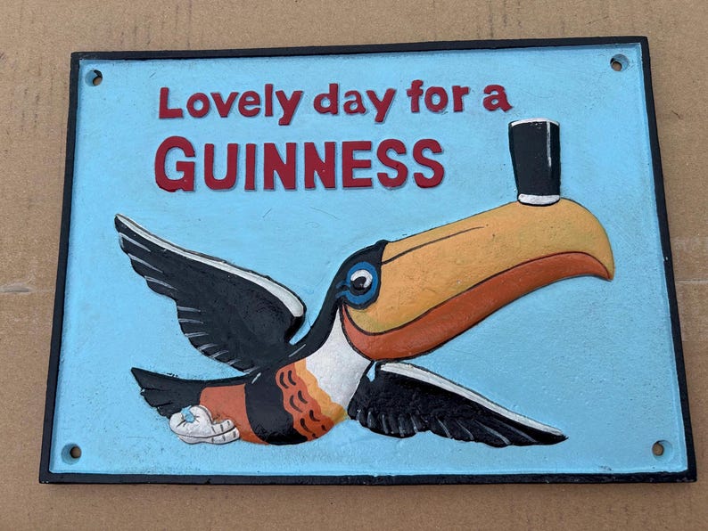 May include: A rectangular metal sign with a light blue background featuring a toucan with a black top hat on its beak. The text reads "Lovely day for a GUINNESS" in red letters. The toucan has black wings, a yellow beak, and a white chest.