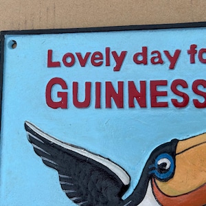 May include: A rectangular metal sign with a light blue background and a black border. The sign features raised red text that reads "Lovely day for a GUINNESS" and a colorful graphic of a toucan with a large yellow and orange beak and black wings.