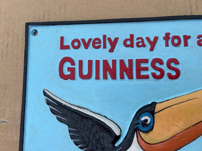 May include: A rectangular metal sign with a light blue background and a black border. The sign features raised red text that reads "Lovely day for a GUINNESS" and a colorful graphic of a toucan with a large yellow and orange beak and black wings.