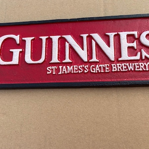 Op de afbeelding: Een rechthoekig bord van gegoten metaal met een rode achtergrond en een zwarte rand. Het woord GUINNESS is in reli&euml;f aangebracht in grote witte letters, met de tekst ST JAMES'S GATE BREWERY DUBLIN in kleinere witte letters eronder.