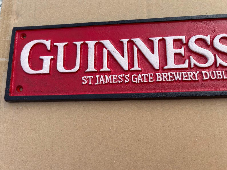 Op de afbeelding: Een rechthoekig bord van gegoten metaal met een rode achtergrond en een zwarte rand. Het woord GUINNESS is in reli&euml;f aangebracht in grote witte letters, met de tekst ST JAMES'S GATE BREWERY DUBLIN in kleinere witte letters eronder.