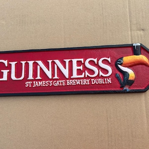 Op de afbeelding: Een rechthoekig gietijzeren bord met een puntig uiteinde, voorzien van een rode achtergrond en een zwarte rand. De tekst GUINNESS en ST JAMES'S GATE BREWERY DUBLIN is in het wit. Rechts staat een kleurrijke illustratie van een toekan.
