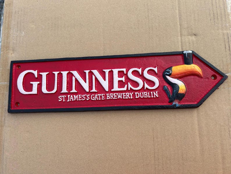 Op de afbeelding: Een rechthoekig gietijzeren bord met een puntig uiteinde, voorzien van een rode achtergrond en een zwarte rand. De tekst GUINNESS en ST JAMES'S GATE BREWERY DUBLIN is in het wit. Rechts staat een kleurrijke illustratie van een toekan.