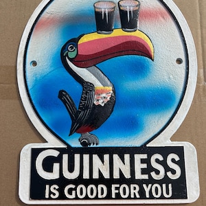 Puede incluir: Un letrero met&aacute;lico circular que presenta un tuc&aacute;n colorido con un pico grande equilibrando dos vasos de cerveza oscura sobre su cabeza. El fondo es un degradado azul y blanco. Debajo del tuc&aacute;n, una base rectangular negra muestra el texto blanco GUINNESS IS GOOD FOR YOU.