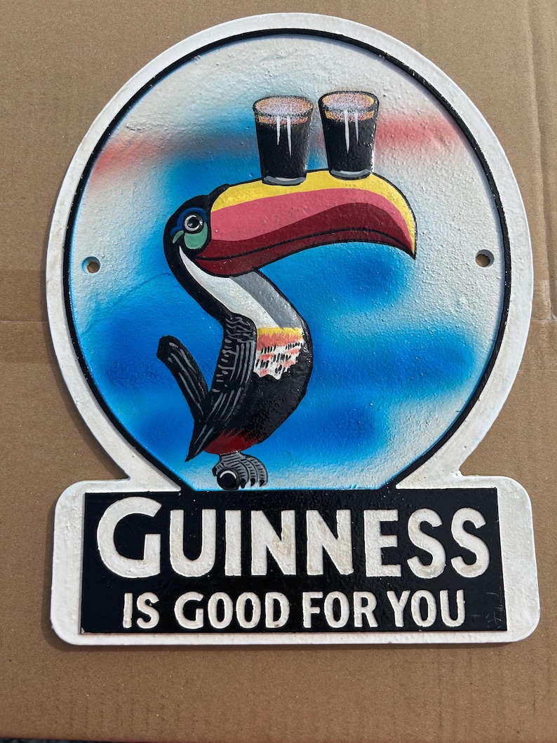 Puede incluir: Un letrero met&aacute;lico circular que presenta un tuc&aacute;n colorido con un pico grande equilibrando dos vasos de cerveza oscura sobre su cabeza. El fondo es un degradado azul y blanco. Debajo del tuc&aacute;n, una base rectangular negra muestra el texto blanco GUINNESS IS GOOD FOR YOU.