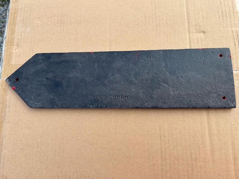 Op de afbeelding: Een rechthoekig zwart metalen bord met een puntig uiteinde, voorzien van de in reli&euml;f aangebrachte tekst 1949 Dublin. Het stuk heeft een getextureerd oppervlak en bevat drie kleine ronde gaten voor montage, met vage rode verfmarkeringen.