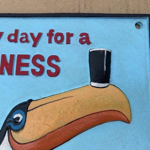 May include: A rectangular metal sign with a light blue background featuring a toucan with a yellow beak balancing a black glass on its tip. The red text reads "lovely day for a GUINNESS" with a black border and a small hole in the top corner.