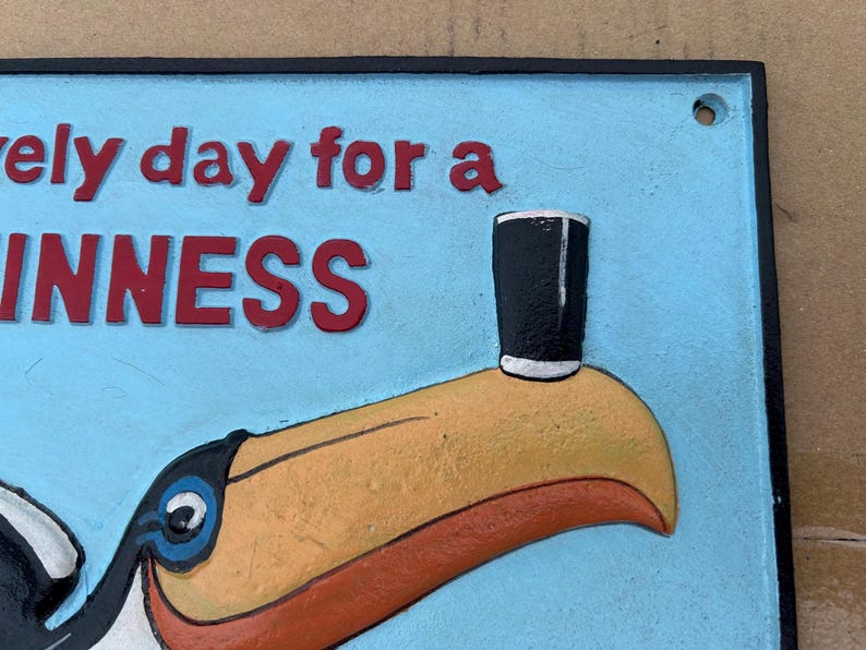 May include: A rectangular metal sign with a light blue background featuring a toucan with a yellow beak balancing a black glass on its tip. The red text reads "lovely day for a GUINNESS" with a black border and a small hole in the top corner.