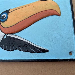 May include: A rectangular metal wall plaque featuring a raised, painted relief of a toucan against a light blue background. The toucan has a large orange and yellow beak, a white neck, and black wings. The plaque has a black border and a small hole.