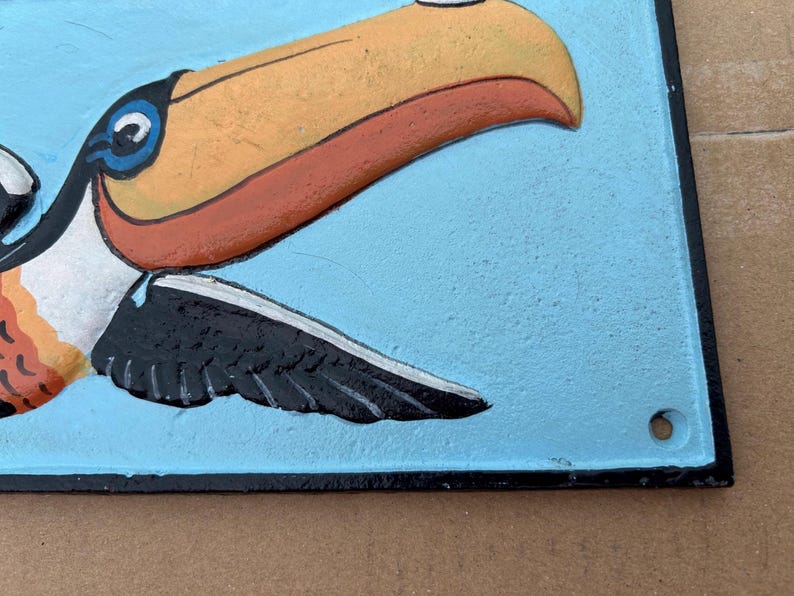 May include: A rectangular metal wall plaque featuring a raised, painted relief of a toucan against a light blue background. The toucan has a large orange and yellow beak, a white neck, and black wings. The plaque has a black border and a small hole.
