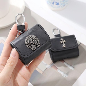 Op de afbeelding: Twee zwarte lederen hoesjes voor draadloze oordopjes, elk voorzien van een zilverkleurig metalen kruisembleem. Het hoesje op de voorgrond heeft een cirkelvormig logo met de tekst CHROME HEARTS en een metalen clip aan de bovenste flap.