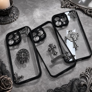 Puede incluir: Tres fundas transparentes para teléfono con bordes negros y aberturas para la cámara. Cada funda presenta emblemas decorativos de metal plateado, incluyendo motivos de cruces y diseños circulares con grabados detallados.