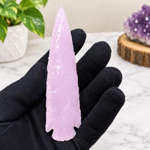 May include: A light pink, translucent, spear-shaped stone point with a sharp, pointed tip and a notched base. The surface of the stone features a smooth, glossy texture with subtle, irregular flaking patterns along the edges of the object.