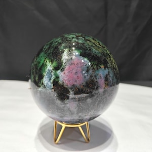 Peut inclure: Une pierre précieuse sphérique polie présentant un motif tacheté de vert forêt profond, de noir et d'inclusions magenta-rose vibrantes. La pierre repose sur un petit support en métal doré à trois pieds, révélant une surface lisse et réfléchissante.