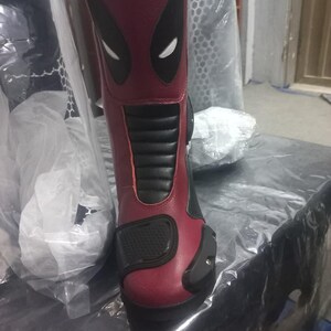 May include: A mid-calf motorcycle boot in deep red and black synthetic leather. The upper shaft features a stylized black and white mask graphic. The boot includes a black ribbed panel on the shin, a textured toe cap, and a reinforced black heel guard.