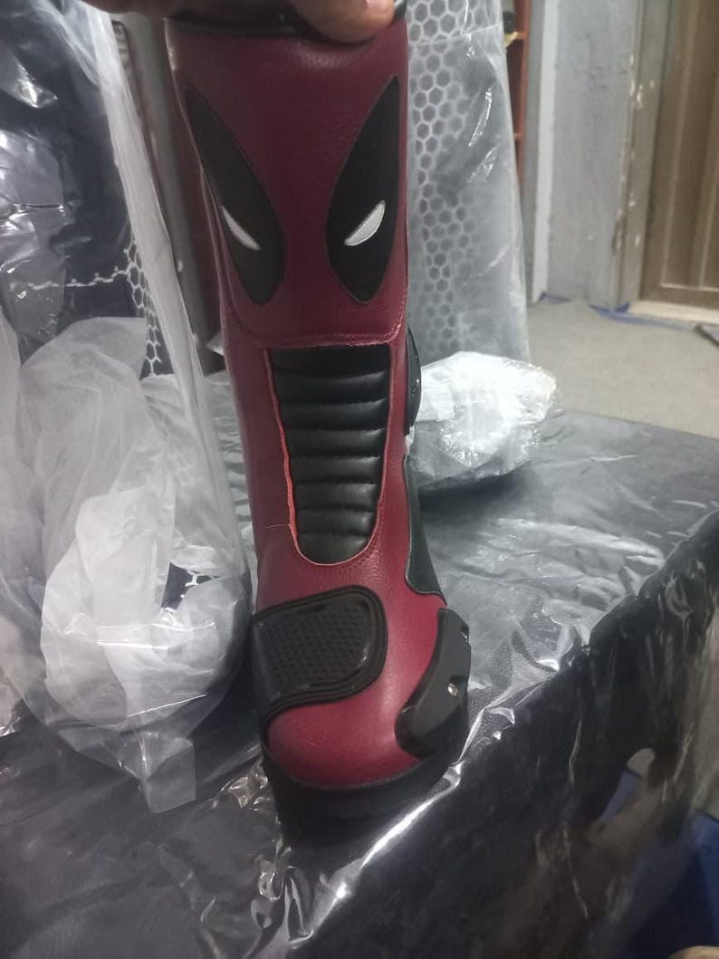 May include: A mid-calf motorcycle boot in deep red and black synthetic leather. The upper shaft features a stylized black and white mask graphic. The boot includes a black ribbed panel on the shin, a textured toe cap, and a reinforced black heel guard.