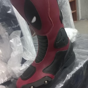 May include: A tall motorcycle boot in deep red and black with a textured finish. The boot features a prominent black oval eye-shaped graphic on the upper shaft, black ribbed panels at the ankle, and a reinforced black toe cap with visible silver screws.
