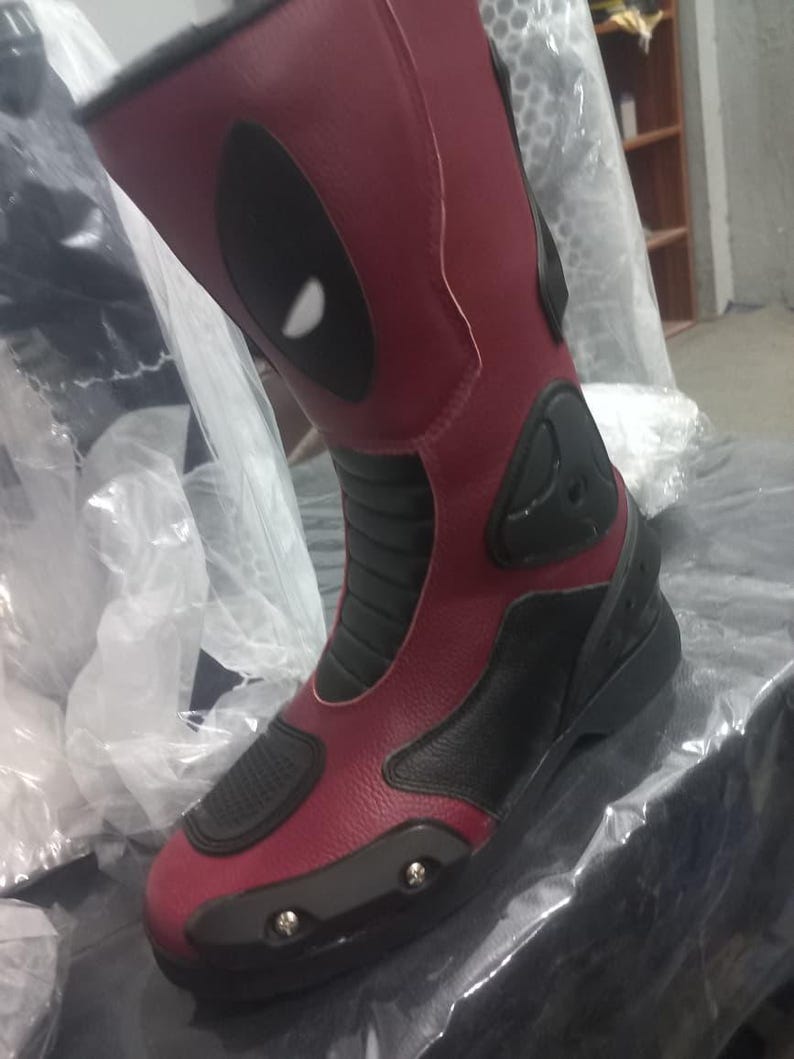 May include: A tall motorcycle boot in deep red and black with a textured finish. The boot features a prominent black oval eye-shaped graphic on the upper shaft, black ribbed panels at the ankle, and a reinforced black toe cap with visible silver screws.