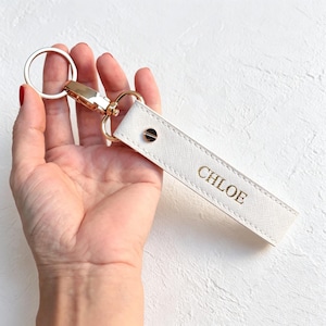 Puede incluir: Una correa rectangular blanca para llavero con superficie texturizada que presenta el nombre CHLOE grabado en letras doradas. La correa está unida a un mosquetón giratorio de metal dorado y a una anilla circular para las llaves.