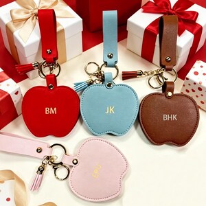 May include: Four apple-shaped keychains with wrist straps, featuring gold-tone hardware and tassels. The red keychain is embossed with BM, the light blue with JK, the brown with BHK, and the light pink with EPU. Each keychain has a gold ring.
