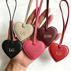 Può includere: Una collezione di etichette per bagagli a forma di cuore in vari colori: nero, beige, rosa, bordeaux e rosso. Ogni etichetta presenta iniziali dorate e un cinturino abbinato. Le etichette sono realizzate in materiale strutturato.