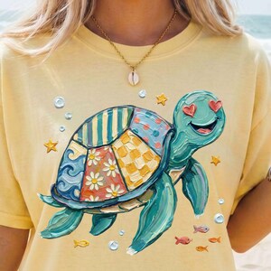 May include: A light yellow t-shirt featuring a colorful turtle design with heart-shaped eyes. The turtle's shell is a patchwork of patterns. The shirt has the text "Ink and Styled" at the bottom.