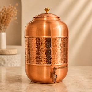 May include: A cylindrical copper water dispenser featuring a hammered texture on the middle section, a smooth finish on the top and bottom, a removable lid with a small gold-toned knob, and a functional metal spigot attached to the lower front.
