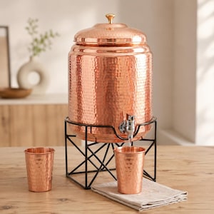 Könnte beinhalten: Ein großer, zylindrischer Wasserspender aus gehämmertem Kupfer mit passendem Deckel und einem silberfarbenen Metallhahn. Der Spender steht auf einem schwarzen Metalldrahtgestell. Zwei passende Becher aus gehämmertem Kupfer stehen daneben.