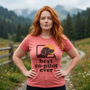 May include: A coral-coloured short-sleeve t-shirt featuring a dark brown graphic of a dog silhouette inside a rounded square frame with a yellow circle behind it. Below the graphic, the text reads "best co-pilot ever" in dark brown letters.