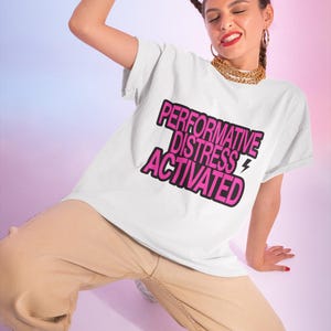 Puede incluir: Una camiseta blanca de manga corta con el texto rosa de borde negro "PERFORMATIVE DISTRESS ACTIVATED" y un pequeño icono de rayo. La prenda tiene cuello redondo y un corte holgado sobre un fondo de color liso.