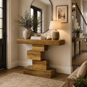 Puede incluir: Una consola de madera con una base geométrica única. La mesa está adornada con un jarrón con vegetación, libros, una vela y una lámpara. Un gran espejo ovalado cuelga de la pared. La habitación tiene una paleta de colores neutros.