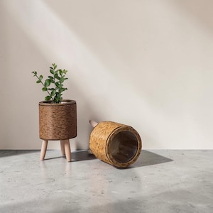 May include: Two cylindrical planters with woven brown textured exteriors and three light-colored wooden legs. One planter stands upright containing a small green leafy plant, while the other lies on its side, revealing a clear interior lining.