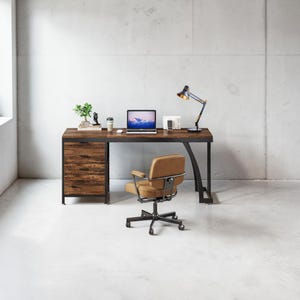 May include: A rectangular desk with a dark wood grain surface and a black metal frame featuring three drawers on the left side. A tan upholstered office chair with a black metal base and wheels is positioned in front of the desk.