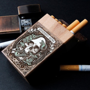 Peut inclure: Un étui à cigarettes en bois clair, orné d'une gravure détaillée d'un crâne entouré de serpents et de l'inscription "MEMENTO MORI". L'étui est ouvert, révélant des cigarettes. Un briquet en bronze est visible en arrière-plan.