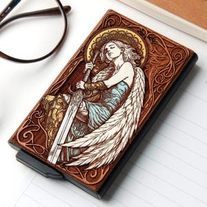 May include: A rectangular card holder featuring a dark wood cover with an intricate laser-engraved illustration of a winged figure holding a large sword. The design includes a golden halo, blue accents on the clothing, and ornate scrollwork borders.