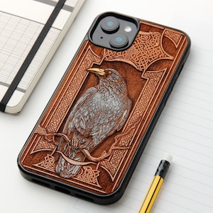 Può includere: Una custodia per smartphone nera con un pannello posteriore color legno caratterizzato da un design inciso al laser di un corvo appollaiato su un ramo, circondato da dettagliati motivi a intreccio celtico che incorniciano l'illustrazione.