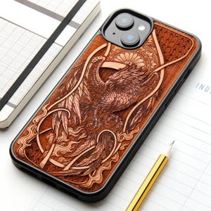 Könnte beinhalten: Eine schwarze Smartphone-Hülle mit einer detaillierten, lasergravierten Holzrückseite. Das Design zeigt einen Phönix mit komplexen Federstrukturen, wirbelnden Flammenmustern und einem kreisförmigen Mandala-Motiv hinter dem Kopf.