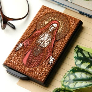 May include: A wooden wallet with a detailed carved design. The design features a skeleton figure in a red robe with a halo, set against a brown background. The wallet has a black edge and a silver-colored clasp.