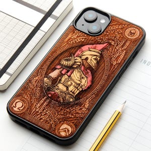 Peut inclure: Une coque de smartphone noire avec une gravure détaillée ton bois représentant un guerrier portant un casque et une armure. Le motif comprend des épis de blé, un emblème circulaire avec un casque et une bordure à motif grec autour du personnage.