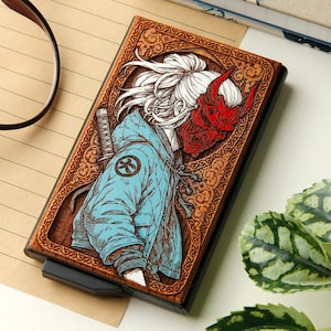 May include: A wooden wallet with a detailed carving of a figure wearing a red demon mask and a blue jacket. The design is framed by an ornate border. The wallet has a black edge and a sliding mechanism.