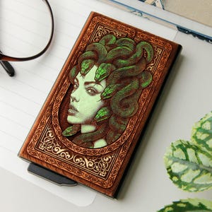 Op de afbeelding: Een bruin en zwart kaarthouder met een illustratie van Medusa, haar haar gevormd door groene slangen. Het kunstwerk is omlijst door een gegraveerde rand. De kaarthouder heeft een modern ontwerp. Afmetingen in centimeters.