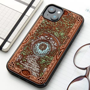 Pode incluir: Uma capa de smartphone preta com um design detalhado gravado em madeira de uma porta redonda inserida numa colina, rodeada por vinhas e folhagem intrincadas. A arte inclui um caminho de pedra que leva à porta, em tons de castanho e azul claro.