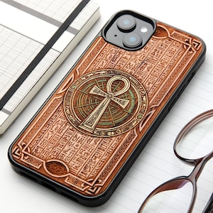 May include: A black smartphone case featuring a wood-toned back panel engraved with intricate Egyptian hieroglyphics and a central circular emblem displaying a golden ankh symbol. The design includes detailed geometric patterns and textures.