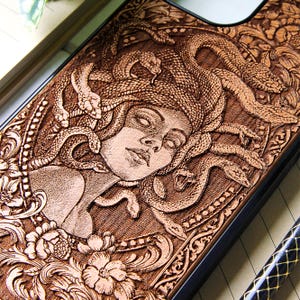 Könnte beinhalten: Braune Handyhülle mit einem eingebrannten Holzbild von Medusa, deren Haare aus Schlangen bestehen. Das Design umfasst florale und verzierte Bordürendetails, die ein klassisches, künstlerisches Gefühl vermitteln. Die Hülle hat einen dunklen Rand.