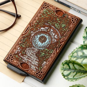 May include: A rectangular card holder featuring a laser-engraved wooden cover with a detailed scene of a circular door set into a stone wall, surrounded by climbing vines and foliage, with light blue accents on the door and green on the leaves.