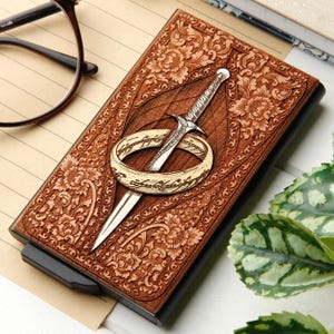 May include: A brown leather wallet with a detailed floral pattern and a silver sword and ring design. The wallet has a black edge and a card slot. The design is inspired by fantasy literature.